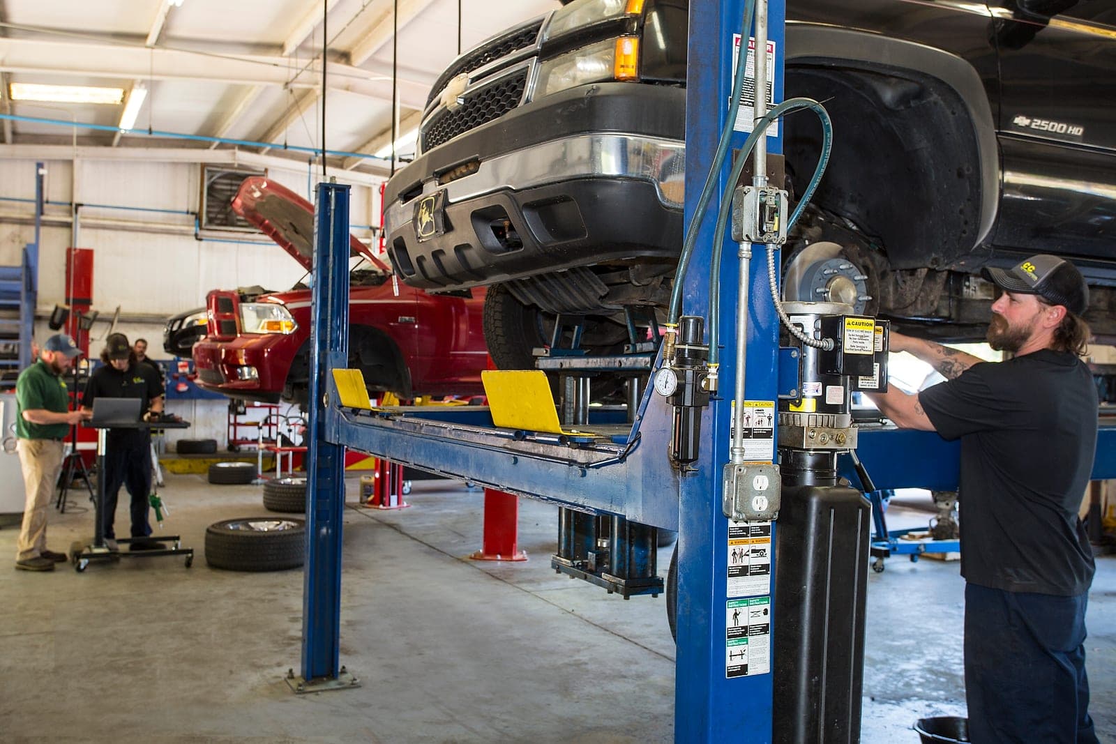 Inside the Warrenville shop showing two vehicles raised on lifts and a technician working on the front suspension of a Chevrolet pickup.
